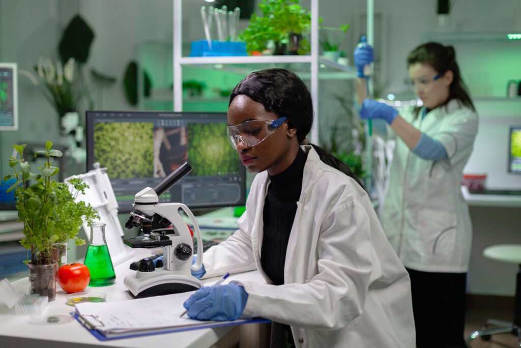 An science woman working in a lab looking into a microscope. At the background there's another person taking a look into a test tube